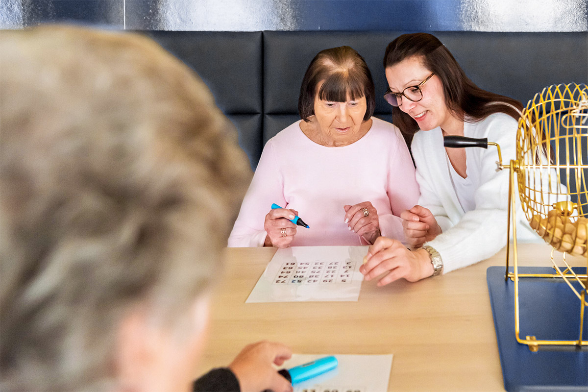 Pflegerin unterstützt Seniorin beim Bingo in einer Tagespflegeeinrichtung